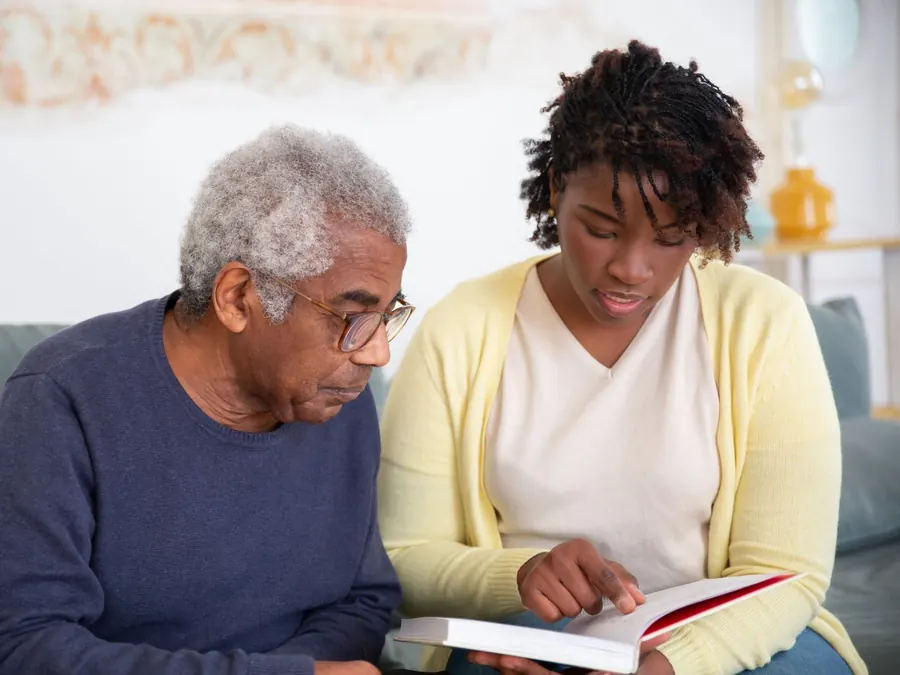 A caregiver and an older man read together on the sofa in his home