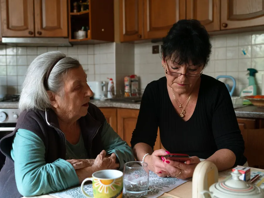 A caregiver and an older woman chat over coffee at her kitchen table