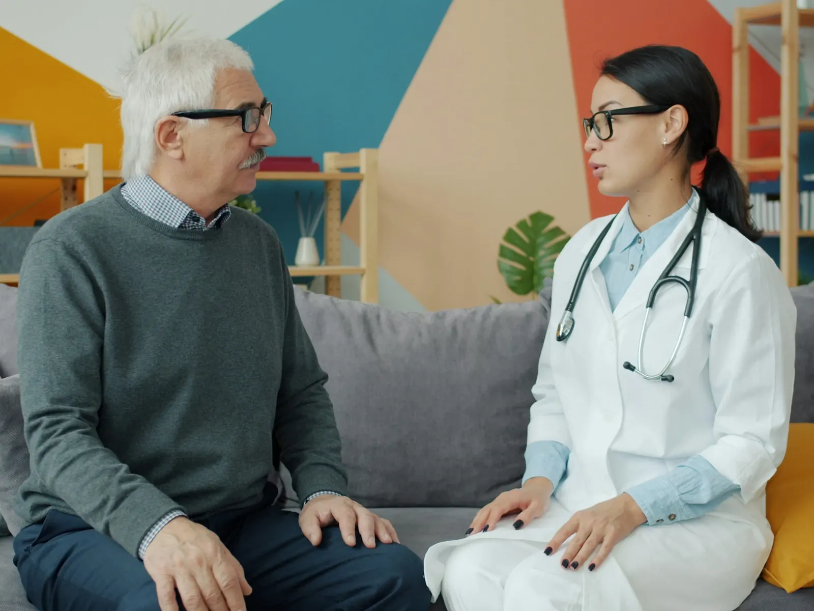 A visiting clinician in a white coat talks with an older man on the couch in his bright living room