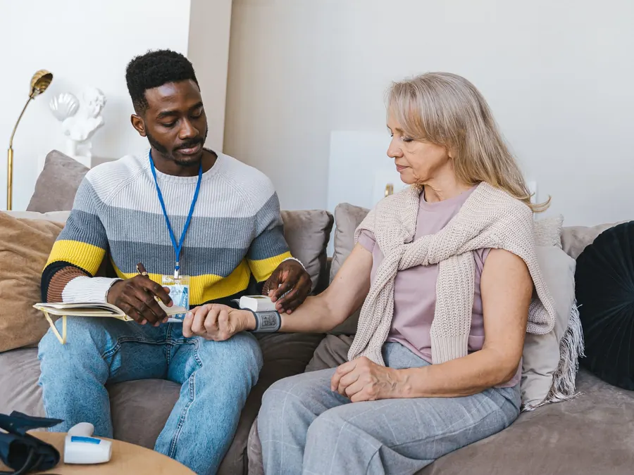 A nurse checks a woman's blood pressure as they sit together on her living room couch