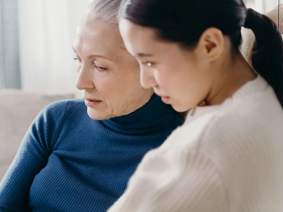A younger woman and an older woman share a quiet conversation on a sofa at home