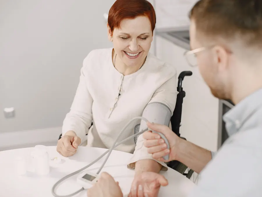 A caregiver checks a smiling woman's blood pressure at her kitchen table